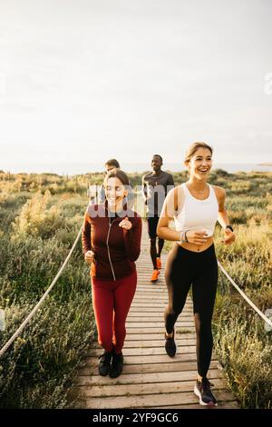 Un groupe d'amis joyeux jogging sur un chemin en bois entouré de verdure luxuriante. Le soleil couchant ajoute de la chaleur, mettant l'accent sur la santé, la forme physique et l'ami Banque D'Images