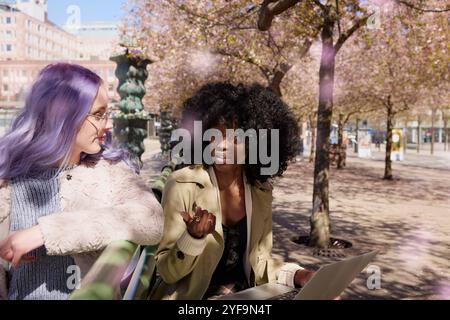 Femme aux cheveux bouclés parlant avec une amie en étant assise sur le banc au parc Banque D'Images