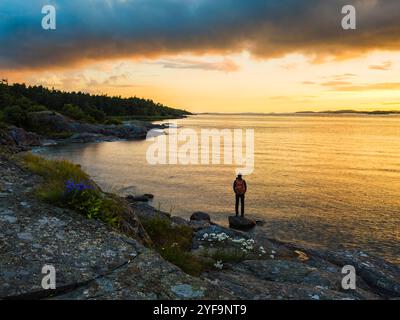Vue arrière de l'homme debout sur le rocher tout en regardant la vue près de la mer sous le ciel sombre pendant le coucher du soleil Banque D'Images