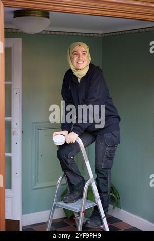 Femme heureuse portant le hijab tenant le détecteur de fumée tout en grimpant l'échelle à la maison Banque D'Images