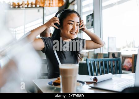 Jeune femme souriante portant des écouteurs tout en étant assise dans un café Banque D'Images