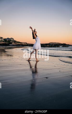 Femme en robe blanche étire ses bras sur la plage au coucher du soleil Banque D'Images