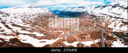 Une vue aérienne à couper le souffle d'une route sinueuse à travers une large vallée en Islande, entourée de montagnes enneigées, d'un terrain rocheux et d'une rivière qui coule Banque D'Images