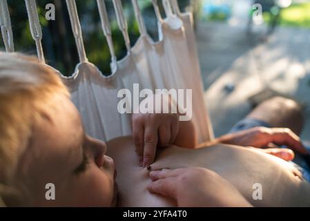 Un garçon de 3 ans regarde un insecte sur sa poitrine tout en étant allongé dans un hamac. Banque D'Images