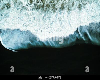 Vue aérienne des vagues qui s'écrasent sur la plage de sable noir d'Islande, créant un contraste saisissant entre le sable sombre et la mousse blanche, capturant le powe brut Banque D'Images