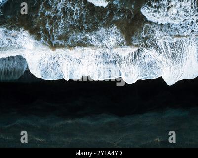 Vue aérienne des vagues qui s'écrasent sur la plage de sable noir d'Islande, créant un contraste saisissant entre le sable sombre et la mousse blanche, capturant le powe brut Banque D'Images