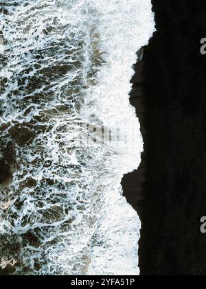 Vue aérienne des vagues qui s'écrasent sur la plage de sable noir d'Islande, créant un contraste saisissant entre le sable sombre et la mousse blanche, capturant le powe brut Banque D'Images