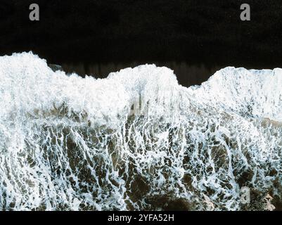 Vue aérienne des vagues qui s'écrasent sur la plage de sable noir d'Islande, créant un contraste saisissant entre le sable sombre et la mousse blanche, capturant le powe brut Banque D'Images