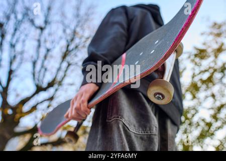 Skateboarder dans un Jean baggy tenant un skateboard rayé marchant dans la rue de la ville. Adolescent fait du skateboard. Concept de sous-culture de la jeunesse Banque D'Images