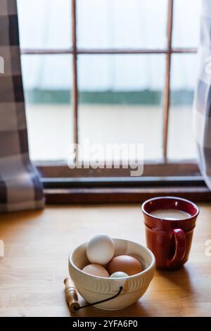 Bol d'oeufs et une tasse rouge sur une table près d'une fenêtre Banque D'Images