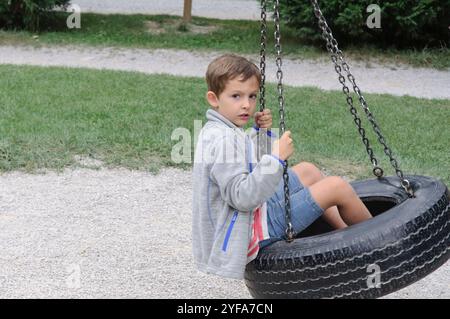 Un jeune garçon assis sur une balançoire de pneu dans une aire de jeux, regardant curieusement la caméra. L'arrière-plan présente une zone herbeuse et un chemin. Banque D'Images