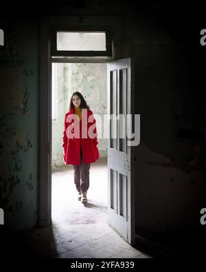 Jeune femme belle vêtue de rouge marchant à travers la porte ouverte d'une chambre abandonnée Banque D'Images