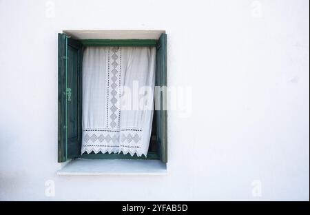 Beau rideau traditionnel blanc ondulant à travers une fenêtre verte en bois ouverte sur un mur blanc Banque D'Images