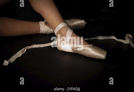 Belles jambes de la jeune ballerine qui met des chaussures pointe au fond noir de sol. Pratique du ballet. Beaux pieds minces gracieux de danseuse de ballet Banque D'Images