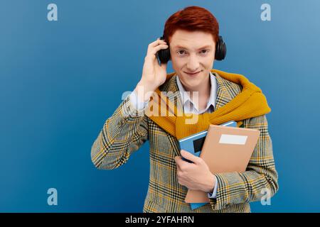 Un jeune homme confiant aux cheveux roux porte une élégante tenue superposée et sourit tout en tenant des livres et des écouteurs. Banque D'Images