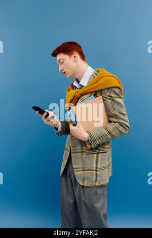 Un jeune homme portant un blazer à carreaux et un pull jaune est concentré sur son téléphone tout en tenant des livres. Banque D'Images