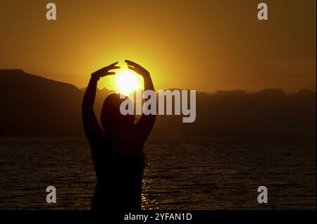 Silhouette de jeune fille à la mer en profitant du coucher du soleil. Lumière du soleil dans l'océan derrière les montagnes Banque D'Images