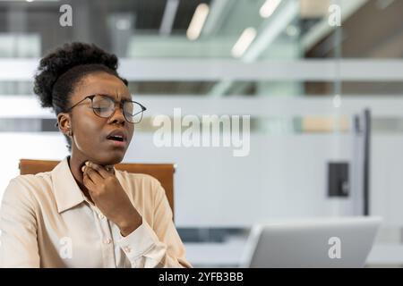 Jeune femme d'affaires afro-américaine se sentant inconfortable dans la gorge au bureau avec ordinateur portable. Semble stressé ou malsain, peut-être dû à des allergies ou au rhume. Thème santé et travail. Banque D'Images