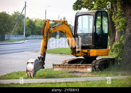 Mini pelle excavatrice orange à chenilles près de la route verte avec arbres sur fond avec Copyspace. Banque D'Images