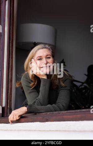 Femme âgée souriante appuyée sur le rebord de la fenêtre, profitant d'un moment paisible à la maison Banque D'Images