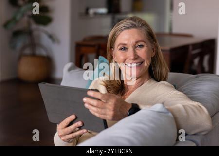 Femme âgée souriante se relaxant à la maison en utilisant une tablette sur un canapé confortable Banque D'Images