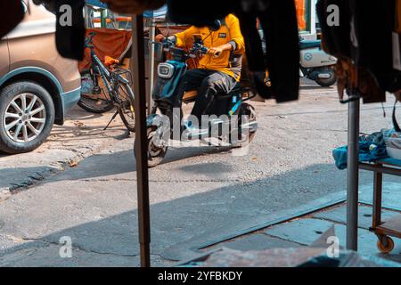 Un livreur de nourriture sur un scooter électrique naviguant dans une rue animée, avec des éléments urbains environnants comme des voitures garées, des vélos Banque D'Images