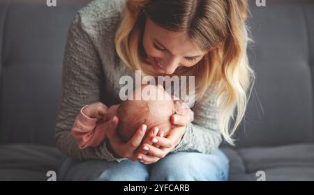 Gros plan mère heureuse tenant tendrement et embrassant le petit bébé, regardant avec amour tout en étant assis sur le canapé gris foncé à la maison. Maman avec nouveau-né à l'intérieur Banque D'Images