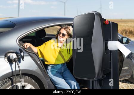 Femme avec des poils courbes portant une chemise jaune s'assoit dans la voiture électrique de charge. Éoliennes en arrière-plan. Banque D'Images