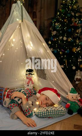 Enfant dans un chapeau de Père Noël dort couché dans un wigwam, à côté d'un arbre de Noël décoré de guirlandes. En attendant un miracle, un conte de fées à la maison. Christma Banque D'Images