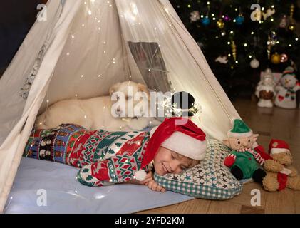 Enfant en chapeau de Père Noël et chien blanc caché dans un wigwam, à côté d'un arbre de Noël décoré. En attendant un miracle, un conte de fées à la maison. Veille de Noël. holi Banque D'Images