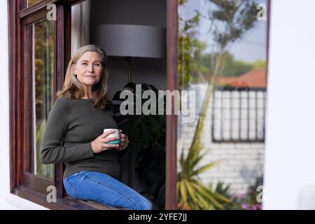 Détente à la maison, femme senior buvant du café tout en regardant par la fenêtre Banque D'Images