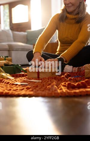 Femme senior souriante enveloppant des cadeaux de Noël sur le sol confortable du salon, à la maison Banque D'Images