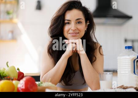 La femme sourit tout en s'appuyant sur son comptoir de cuisine avec un verre de lait et de légumes frais Banque D'Images