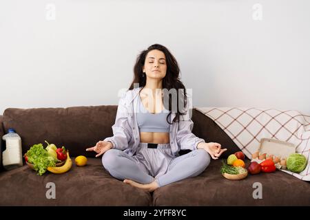 Jeune femme pratiquant le yoga à la maison sur le canapé, méditant les yeux fermés entourée de récipients remplis d'aliments sains Banque D'Images