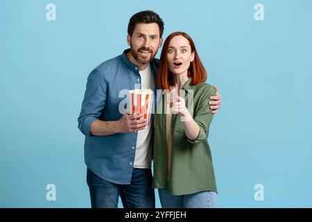 Jeune couple tient un seau de pop-corn, la femme pointe son doigt et ils ont tous les deux l'air surpris Banque D'Images