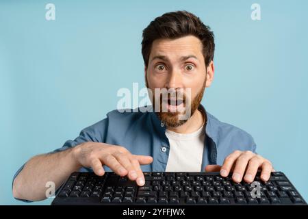 Homme choqué avec les yeux grands ouverts regardant la caméra tout en tapant sur le clavier de l'ordinateur sur fond bleu Banque D'Images