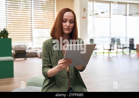 Jeune femme d'affaires est assise dans le hall de son bureau, se concentrant sur la lecture de quelque chose sur sa tablette Banque D'Images