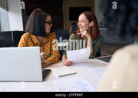 Deux femmes d’affaires partagent un rire tout en analysant des graphiques et des données lors d’une réunion productive au bureau Banque D'Images