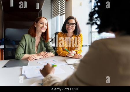 Trois femmes d'affaires sont assises à une table avec des graphiques et des documents, souriant tout en ayant une réunion productive Banque D'Images