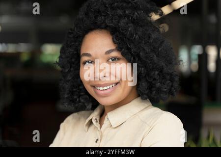 Une jeune femme noire aux cheveux bouclés sourit les bras pliés dans un bureau Banque D'Images