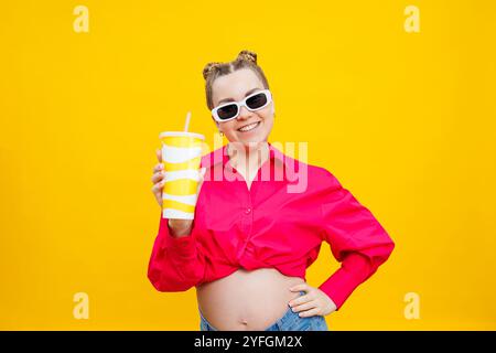 Femme enceinte joyeuse en chemise rose tenant une boisson savoureuse sur fond jaune isolé. Une boisson rafraîchissante pendant la grossesse. Une femme enceinte boit Banque D'Images