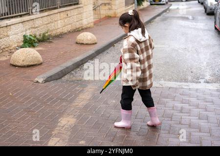 Une jeune fille se promène le long d'une rue humide, vêtue d'une veste à carreaux confortable et de bottes de pluie roses. Elle tient un parapluie coloré, profitant de la bruine tout en explorant son quartier. Banque D'Images