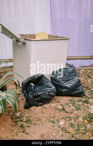 Une poubelle avec deux sacs poubelles noirs placés à côté, prêts à être éliminés dans un espace extérieur Banque D'Images