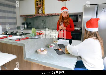 Deux femmes portant des chapeaux de père noël appréciant le dîner de noël avec des sushis et du vin dans une cuisine moderne Banque D'Images