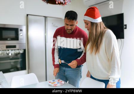 Homme hispanique et femme portant un chapeau de père noël regardant un morceau de gâteau de noël dans leur cuisine moderne Banque D'Images