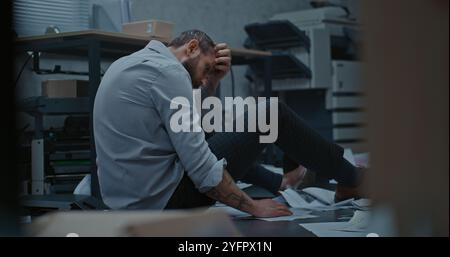 Homme d'affaires déprimé s'assoit sur le sol après la crise de rage sur l'affaire infructueuse. L'employé de bureau bouleversé respire fortement après avoir détruit la salle de bureau. Crise financière ou licenciement. Épuisement du travail. Banque D'Images