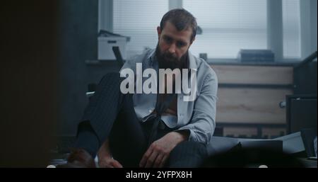 Homme d'affaires déprimé s'assoit sur le sol après la crise de rage sur l'affaire infructueuse. L'employé de bureau bouleversé respire fortement après avoir détruit la salle de bureau. Crise financière ou licenciement. Épuisement du travail. Banque D'Images