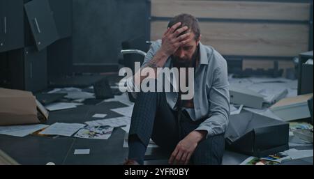 L'employé nerveux et stressé respire fortement, s'assoit sur le sol après avoir détruit la salle de bureau en raison d'un tir. Homme d'affaires frustré après l'échec d'un accord commercial ou recevoir de mauvaises nouvelles. Crise financière Banque D'Images