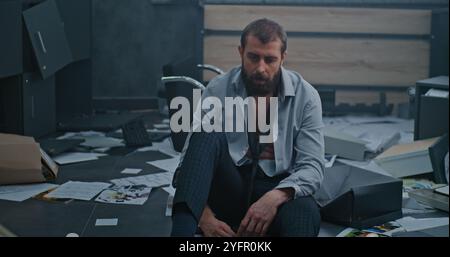 L'employé nerveux et stressé respire fortement, s'assoit sur le sol après avoir détruit la salle de bureau en raison d'un tir. Homme d'affaires frustré après l'échec d'un accord commercial ou recevoir de mauvaises nouvelles. Crise financière Banque D'Images