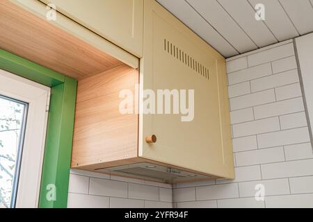 Fragment d'armoires de cuisine supérieures avec façades jaunes et boutons en bois installés sur un mur carrelé autour de la fenêtre Banque D'Images